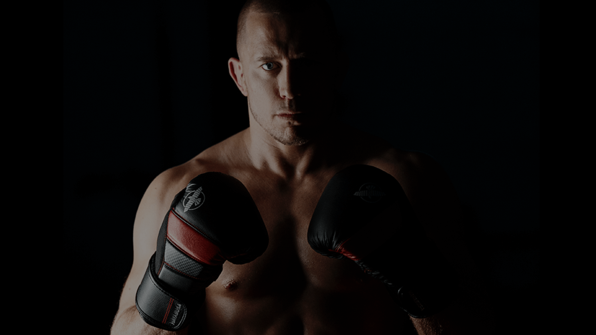 Een gespierde vechtsporter met gekruiste rode bokshandschoenen voor de borst in een donkere omgeving.