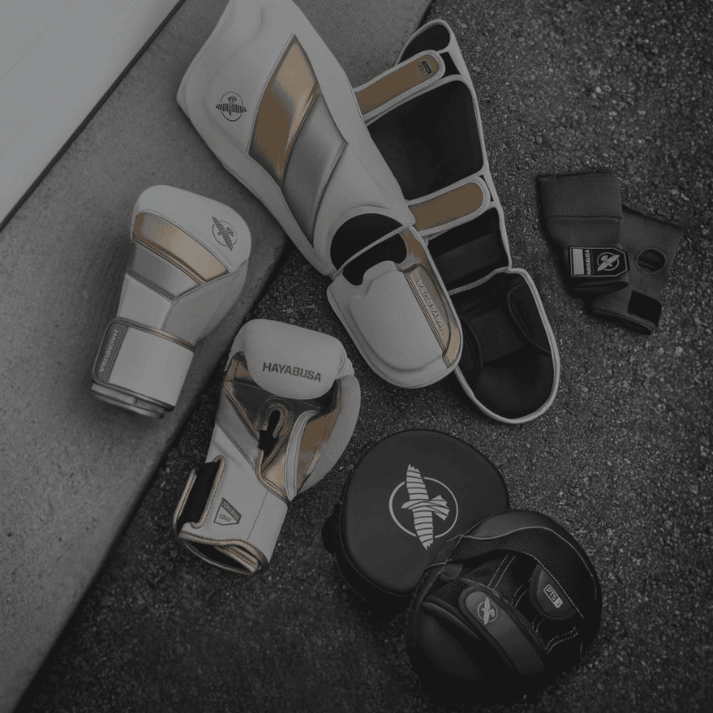 Verschillende vechtsportbeschermers en accessoires, waaronder scheenbeschermers en handwraps, netjes naast elkaar op een grijze ondergrond.