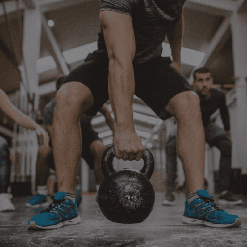 Een persoon in een sportschool doet een kettlebell oefening; we zien de persoon vanuit een laag standpunt, gefocust op de kettlebell en de onderste helft van het lichaam, met sportschoenen aan en een ander persoon op de achtergrond.