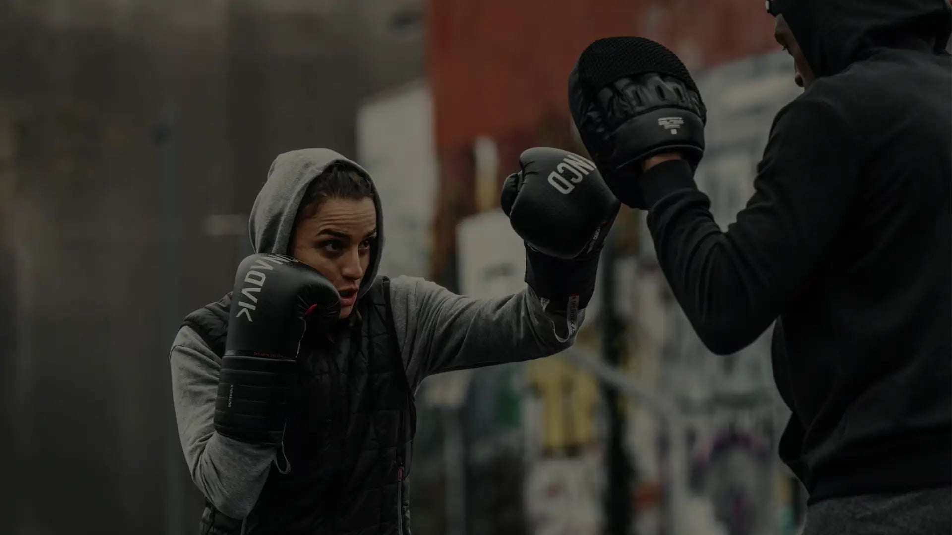 Een sporter traint buiten met zwarte bokshandschoenen en slaat op handpads die worden vastgehouden door een partner in donkere sportkleding.
