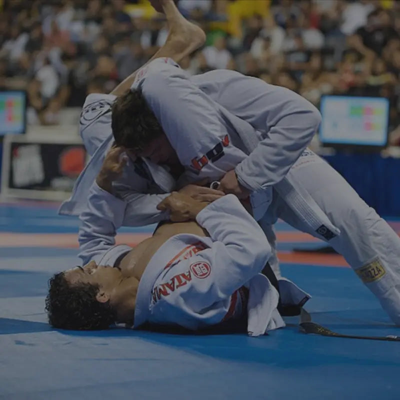 Twee vechters in witte gi’s doen een Braziliaans Jiu Jitsu-wedstrijd op een blauwe mat; één vechter probeert vanaf de rug een armklem toe te passen, terwijl de ander verdedigt.