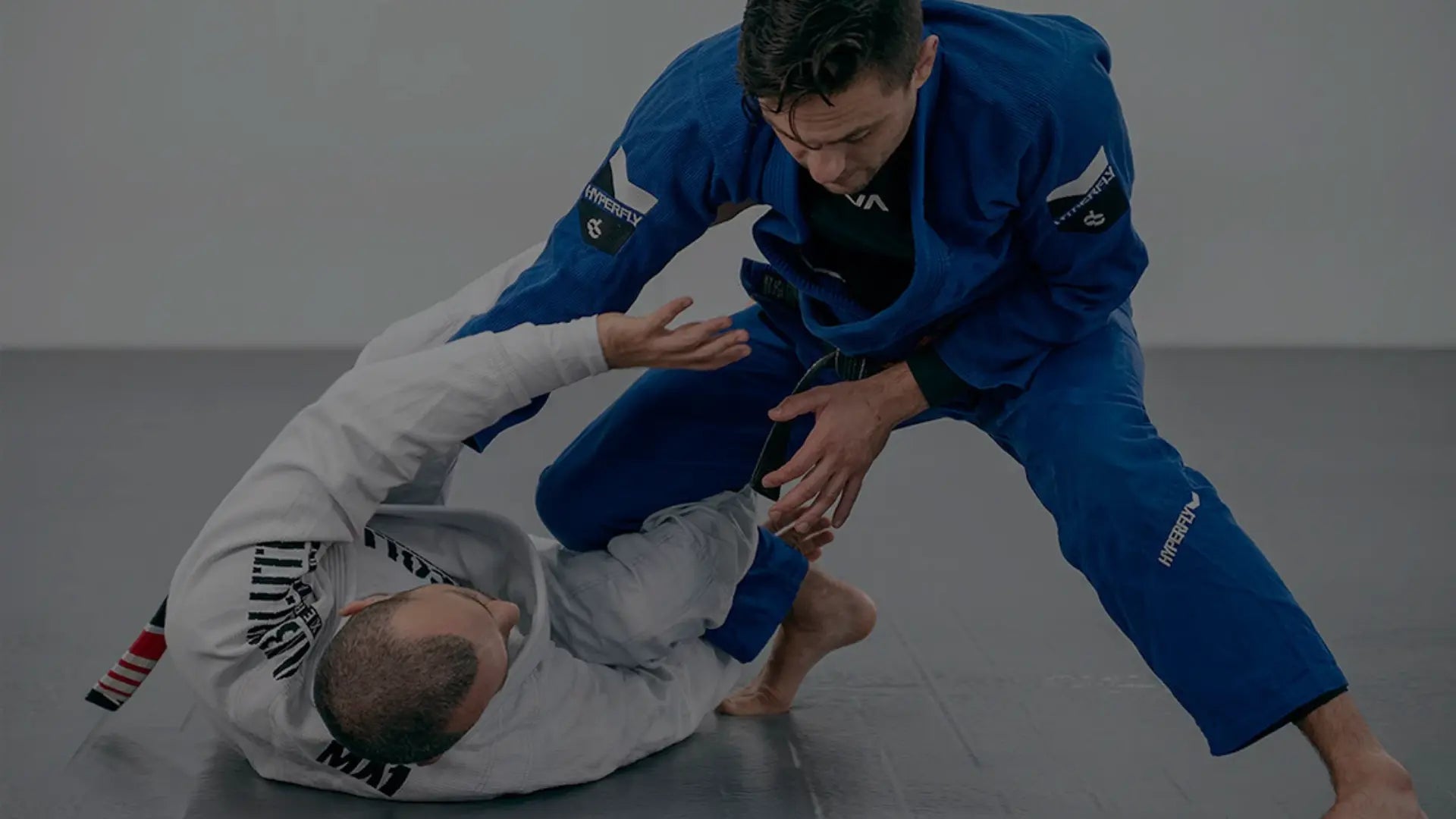 Twee vechters in witte gi’s doen een Braziliaans Jiu Jitsu-wedstrijd op een blauwe mat; één vechter probeert vanaf de rug een armklem toe te passen, terwijl de ander verdedigt.