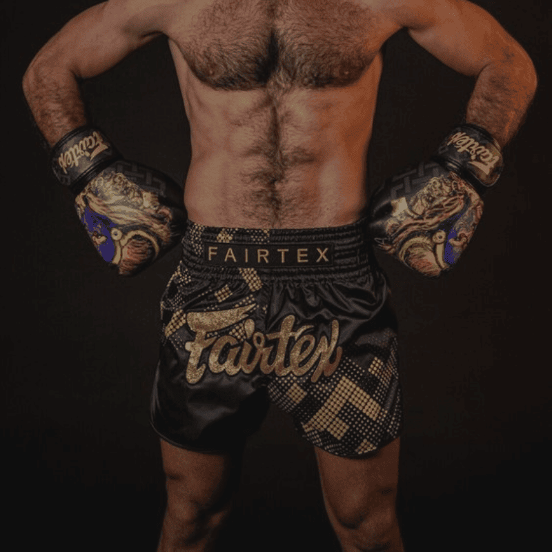 Een getrainde mannelijke vechtsporter van achteren gezien, met Fairtex bokshandschoenen en bijpassende shorts, poseert met gebalde vuisten en opgeheven armen om zijn gespierde rug en schouders te tonen.