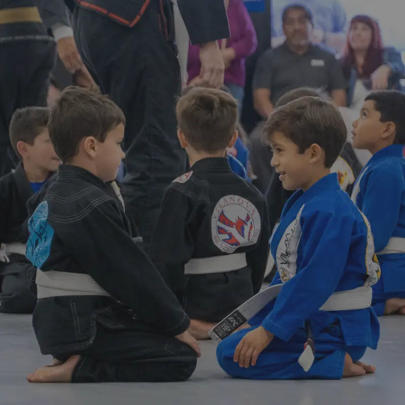 Twee jonge jongens in jiu-jitsu gi's met witte banden zitten tegenover elkaar op de mat en lachen tijdens een kindermartial arts-les, omringd door andere kinderen in zwarte en blauwe gi's.
