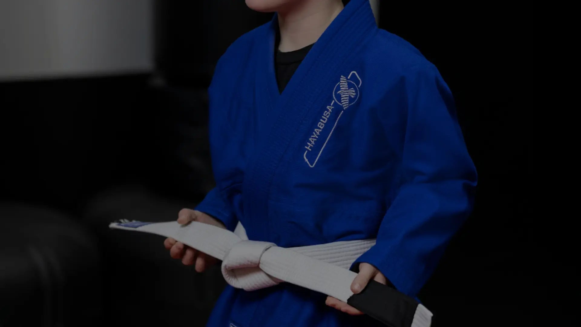 Twee jonge jongens in jiu-jitsu gi's met witte banden zitten tegenover elkaar op de mat en lachen tijdens een kindermartial arts-les, omringd door andere kinderen in zwarte en blauwe gi's.