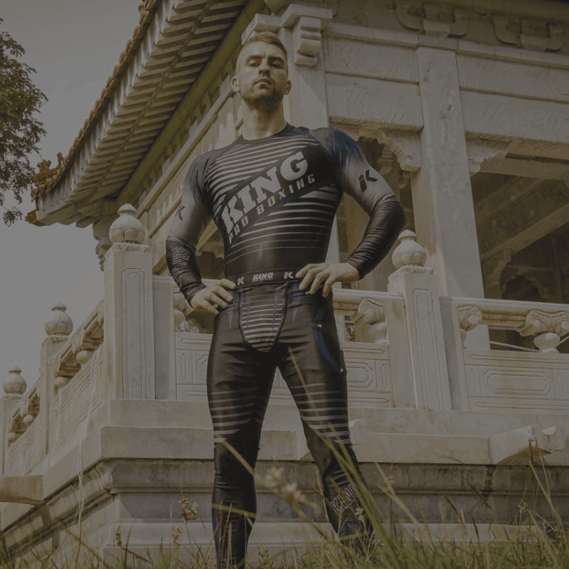 Een man staat zelfverzekerd op een trap voor een tempel, gekleed in een getailleerde legging en een sportshirt met de tekst "KING".