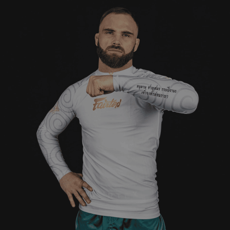 Een man met een getrimde baard en korte haar, gekleed in een witte lange mouwen Fairtex rashguard met Fairtex-logo op de borst en Thaise schrift op de mouwen, poseert met gebalde vuist voor een donkere achtergrond.