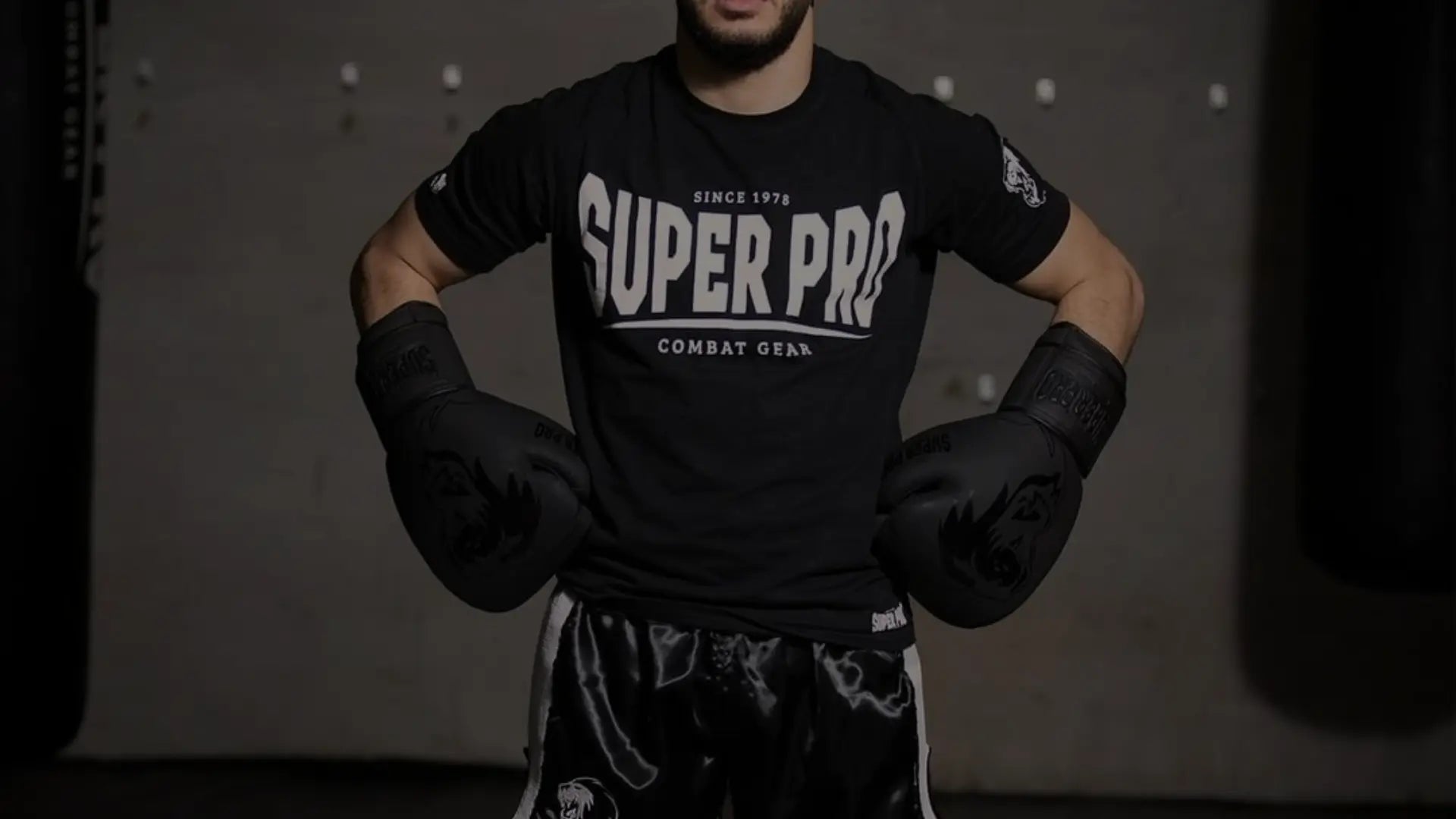 Bokser in een zwarte Super Pro-shirt en zwarte bokshandschoenen staat met de handen in de zij, klaar voor training in een donkere gymomgeving.