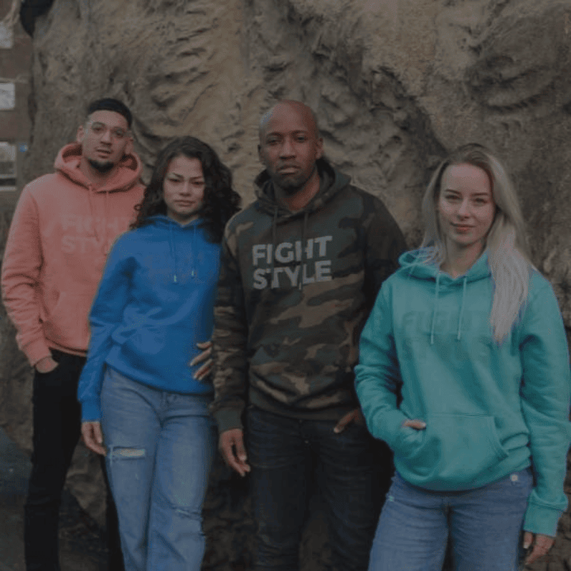 Vier personen, gekleed in 'Fight Style' hoodies in verschillende kleuren, poseren voor een rotsachtige achtergrond.