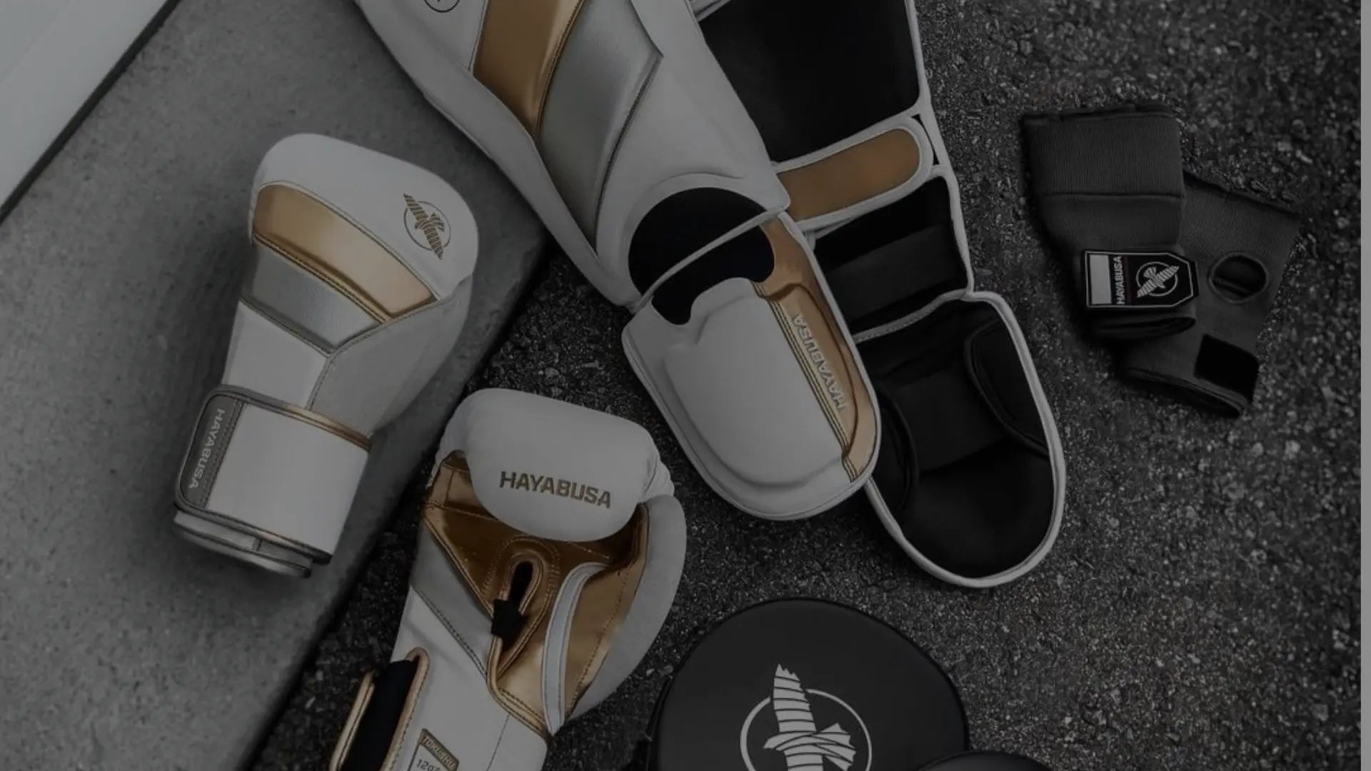 Verschillende vechtsportbeschermers en accessoires, waaronder scheenbeschermers en handwraps, netjes naast elkaar op een grijze ondergrond.