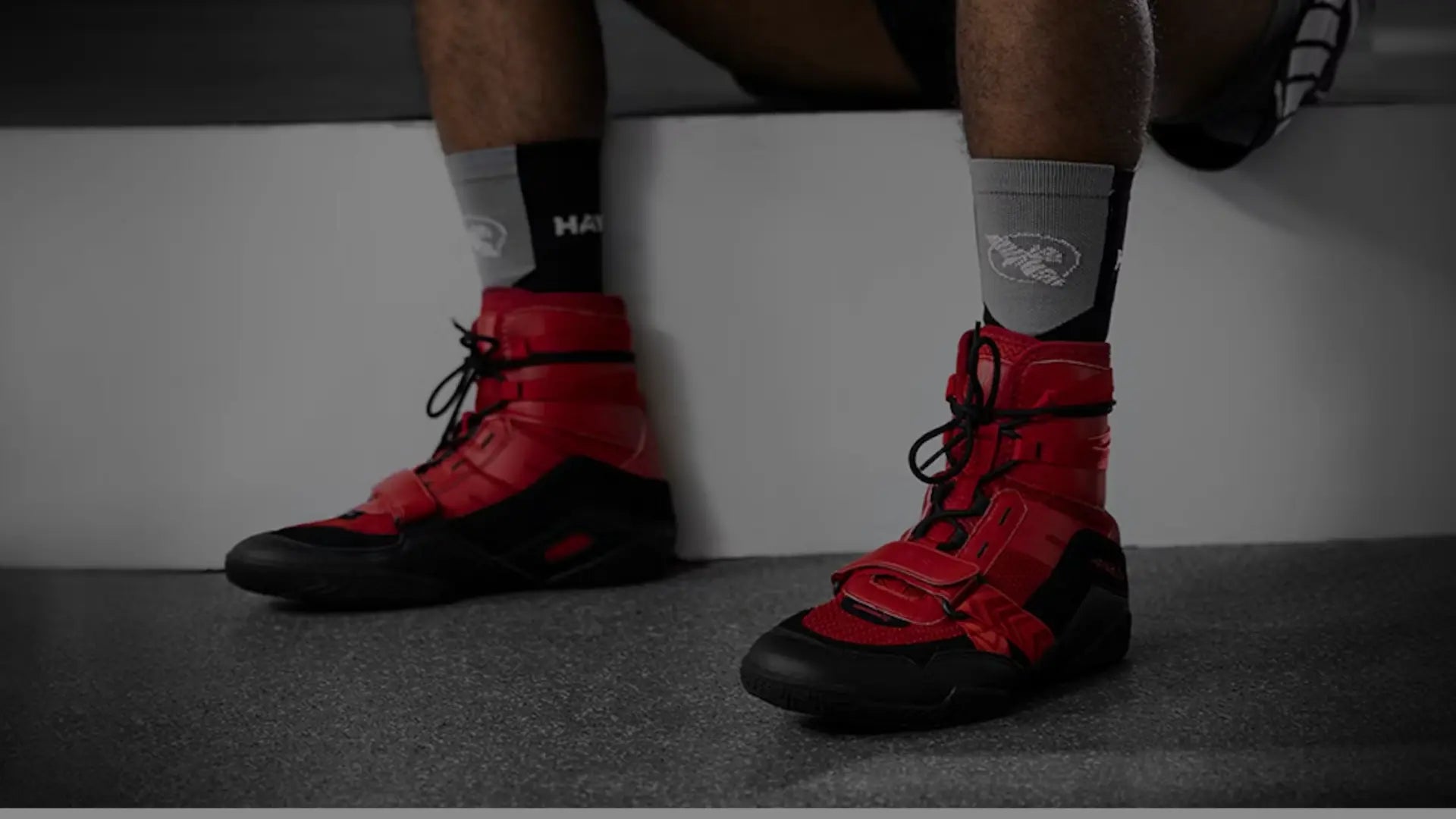 Close-up van een sporter die rode boksschoenen draagt tijdens een training, zittend op een bankje met zicht op de stevige zool en vetersluiting.