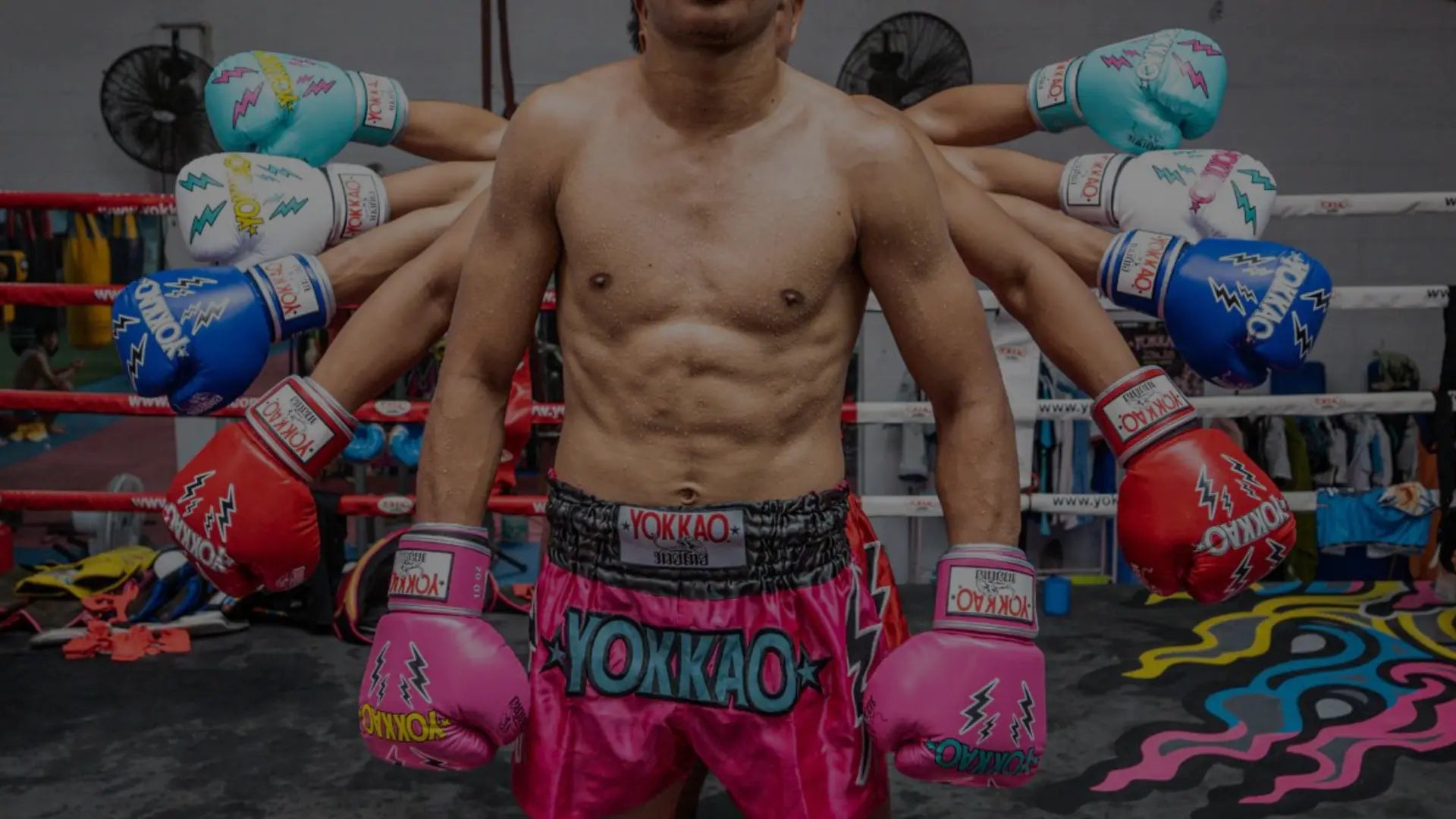 Een man met verschillende kleurrijke bokshandschoenen uitgestrekt op zijn armen in een bokszaal, draagt een roze short