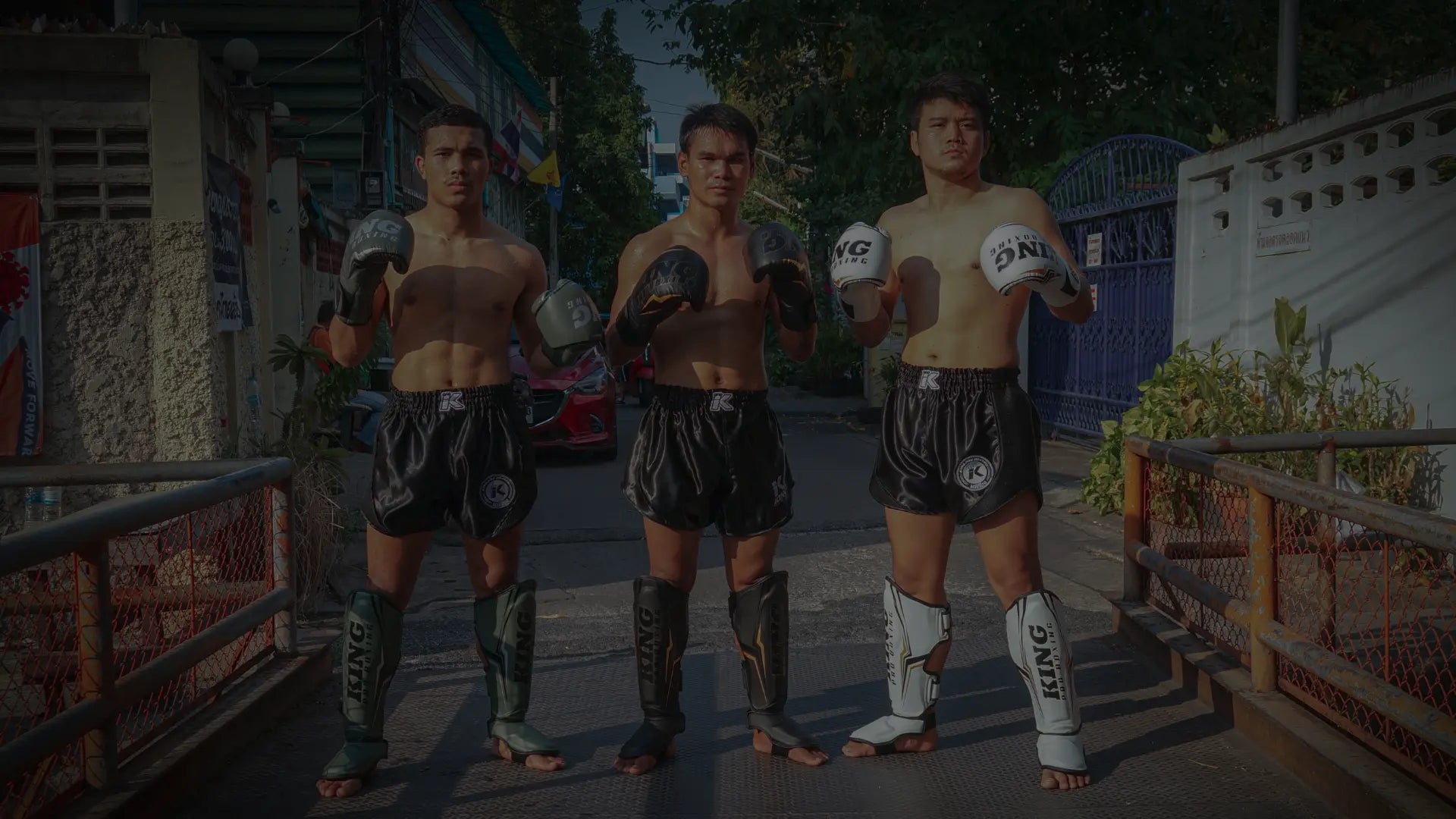Drie mannen in traditionele Muay Thai-houding, gekleed in zwarte shorts en met bokshandschoenen en scheenbeschermers, staan klaar om te trainen op een straat in Thailand.