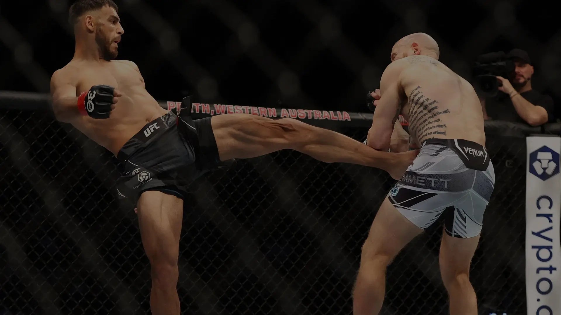 Twee vechters in een kooi waarbij één vechter een hoge trap plaatst naar het lichaam van zijn tegenstander tijdens een intens gevechtsmoment.