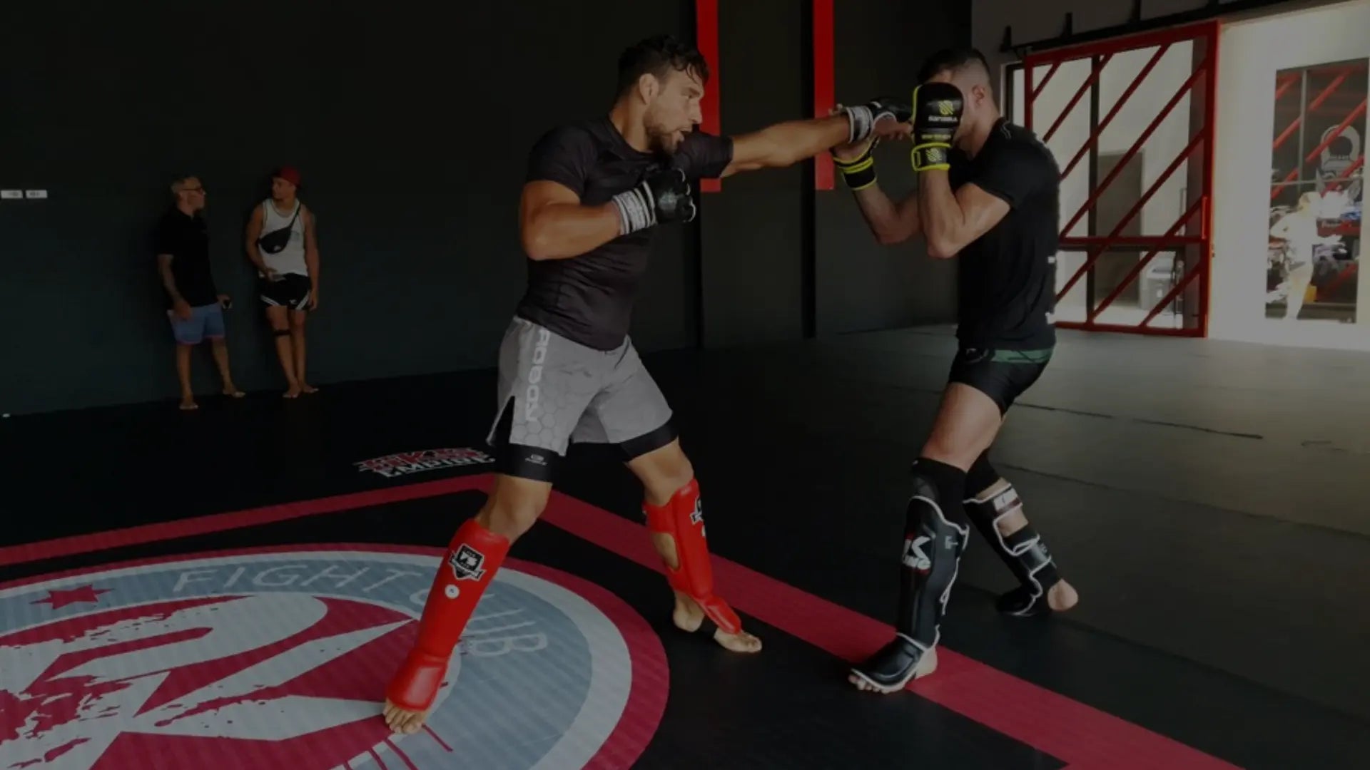Twee kickboksers sparren op de mat, waarbij één vechter een rechte stoot uitdeelt terwijl beide sporters scheenbeschermers en handschoenen dragen tijdens een technische trainingssessie.