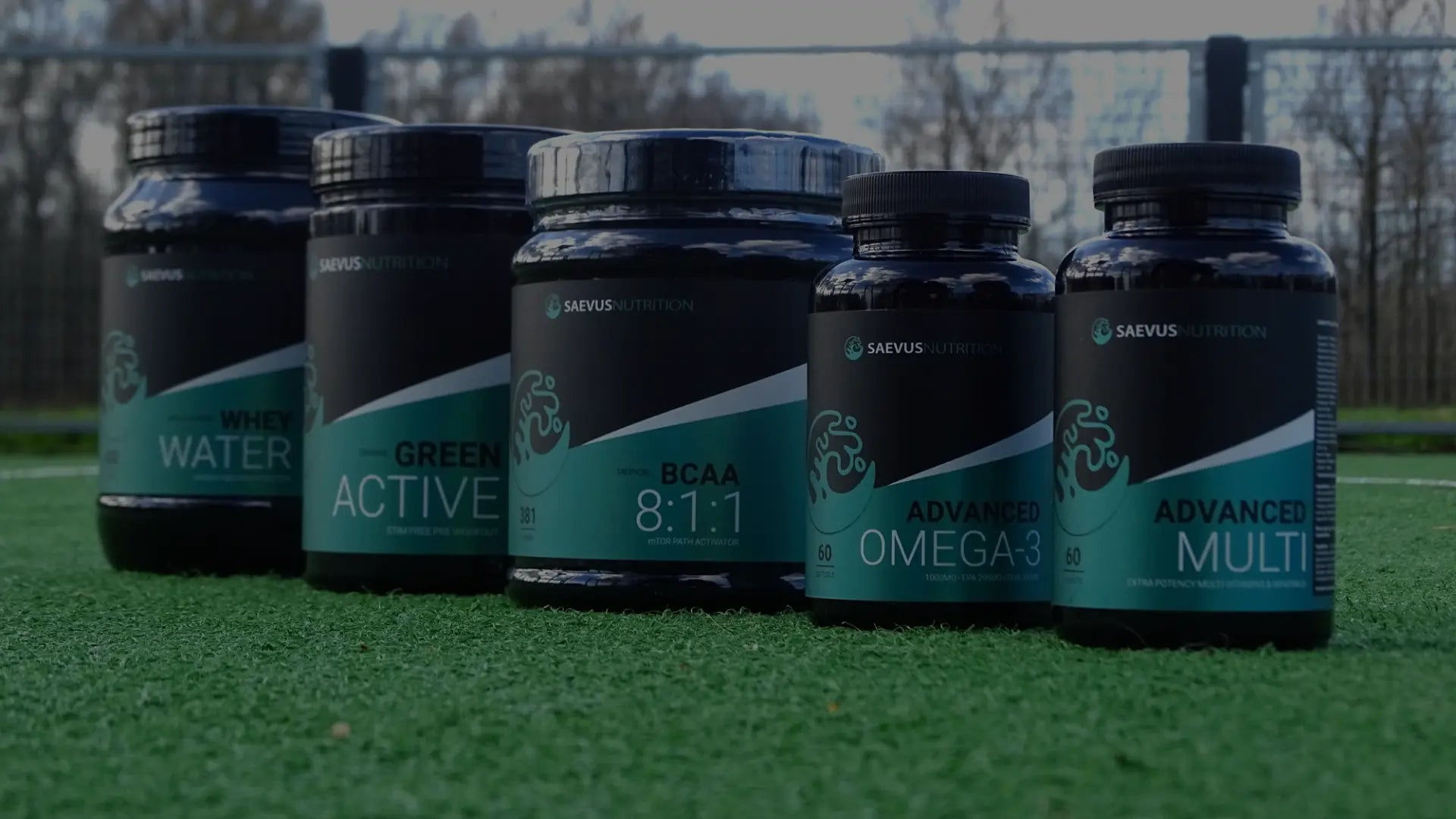 Een rij Saevus Nutrition supplementen – waaronder BCAA, whey water, omega-3 en multivitaminen – uitgestald op een sportveld voor een sportieve en gezonde lifestyle-uitstraling.