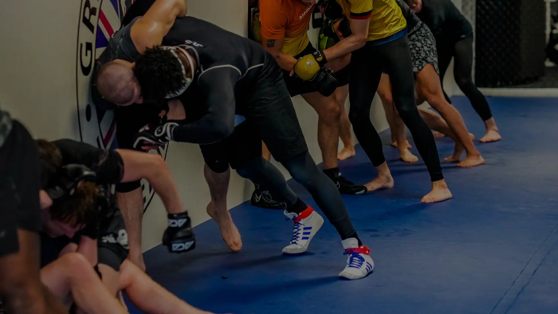 Een groep atleten traint worstel- en grapplingtechnieken tegen de muur, waarbij paren elkaar onder hoge druk controleren en takedowns oefenen tijdens een intensieve gymsessie.