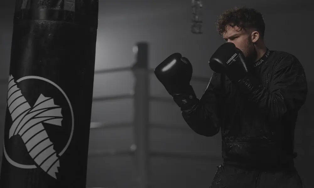 Bokser in donkere trainingsruimte die zwarte bokshandschoenen draagt en een stoot richting een zware bokszak plaatst. De setting is gedimd en geconcentreerd.