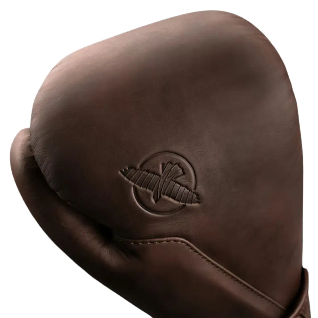 Detailfoto van een bokshandschoen in een warme, diepe bruintint met een matte, luxe afwerking. Het subtiele reliëflogo in dezelfde kleur zorgt voor een minimalistische en stijlvolle uitstraling. De fijne structuur van het materiaal en de strakke stiknaden benadrukken de hoogwaardige kwaliteit en duurzame afwerking van de handschoen.