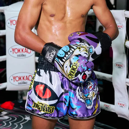 Atleet draagt paarse Yokkao bokshandschoenen met Angry Tiger print en bijpassende Muay Thai shorts met kleurrijke tijgerillustraties. De set heeft een zwarte elastische tailleband met Yokkao-logo en opvallende grafische details. Gefotografeerd in een boksomgeving met Yokkao branding op de achtergrond.