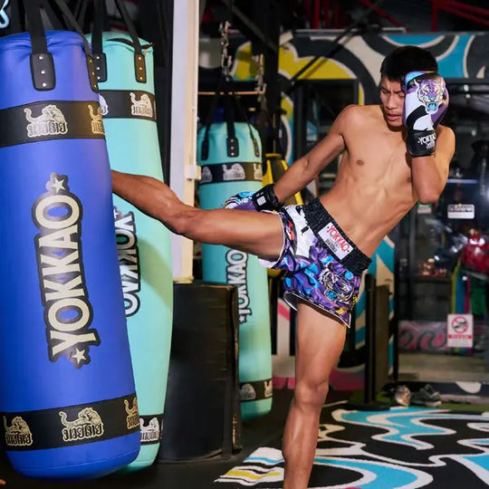 Atleet draagt Yokkao Angry Tiger bokshandschoenen en bijpassende Muay Thai shorts terwijl hij een hoge trap uitvoert tegen een Yokkao bokszak. De paarse set met tijgerprint valt op door het kleurrijke design en zwarte tailleband met Yokkao-logo. De foto is genomen in een gymomgeving met meerdere hangende bokszakken op de achtergrond.