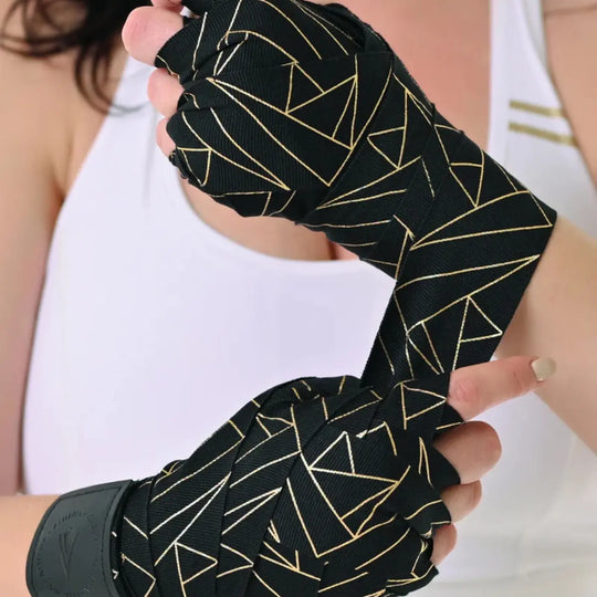 Close-up van de Athena Fightwear handwraps terwijl ze om de handen worden gewikkeld. Het geometrische goud-op-zwart design valt op en biedt een stijlvolle look. De wikkeling toont een stevige en nauwsluitende pasvorm voor boksers. Ideaal voor bescherming en stabiliteit tijdens combinaties.