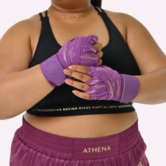 Model draagt de paarse Athena Fightwear Diana Violet handwraps met goudkleurige details. De wraps zitten stevig om beide handen gewikkeld voor optimale ondersteuning tijdens bokstraining. De close-up laat het vrouwelijke, stijlvolle ontwerp mooi uitkomen.