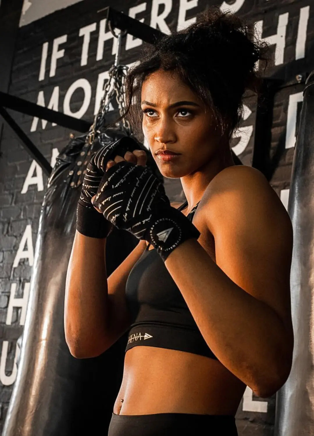 Vrouwelijke sporter in de gym met Athena Fightwear handwraps in zwart, staand in vechthouding bij de bokszak met focus op kracht, grip en ondersteuning.