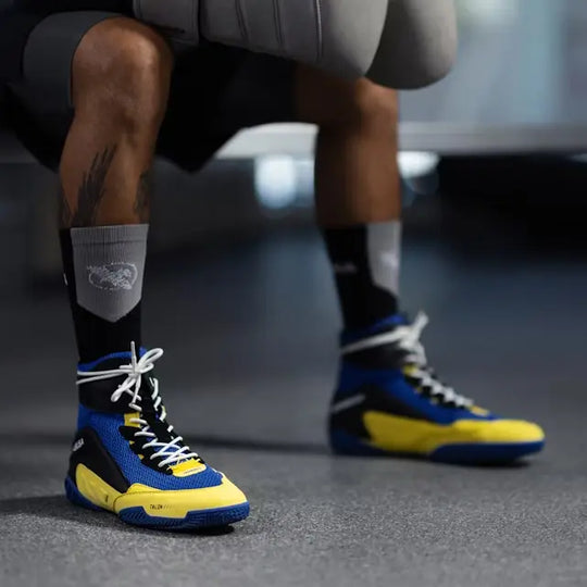 Blauwe en gele boksschoenen van Hayabusa met witte veters, gedragen door een sporter, gefotografeerd op een sportvloer.