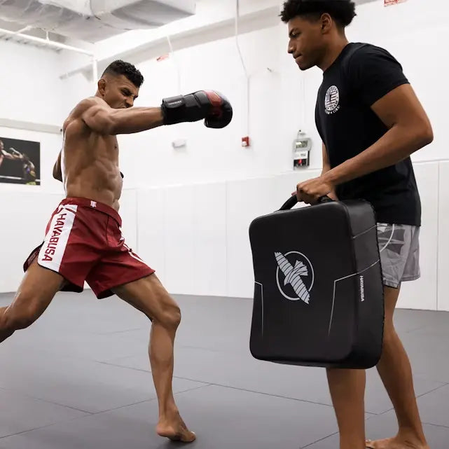 Atleet met rode Hayabusa shorts slaat op een zwarte Hayabusa trainingspad die wordt vastgehouden door een trainingspartner in een witte zaal met lichtgrijze vloer.