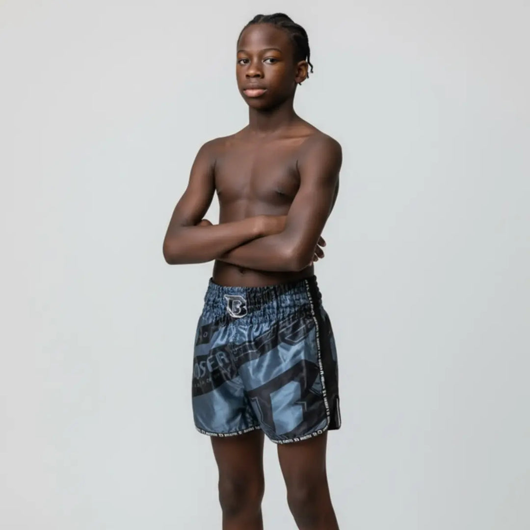 Op deze afbeelding draagt een jonge atleet het Booster TBT Elite 1 Youth kickboksbroekje. De pasvorm, lengte en bewegingsvrijheid van het broekje worden duidelijk zichtbaar. De grijs-zwarte print geeft een stoere, moderne uitstraling tijdens training of sparren.