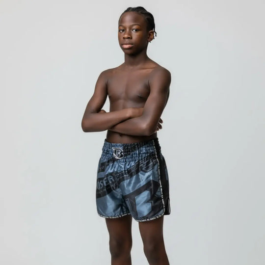 Op deze afbeelding draagt een jonge atleet het Booster TBT Elite 1 Youth kickboksbroekje. De pasvorm, lengte en bewegingsvrijheid van het broekje worden duidelijk zichtbaar. De grijs-zwarte print geeft een stoere, moderne uitstraling tijdens training of sparren.