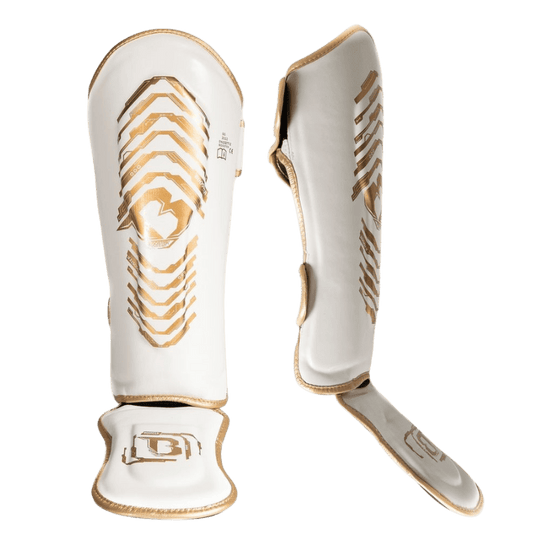 Twee witte scheenbeschermers van het merk Booster, met gouden accenten en voetbescherming, getoond in een voor- en zijaanzicht.