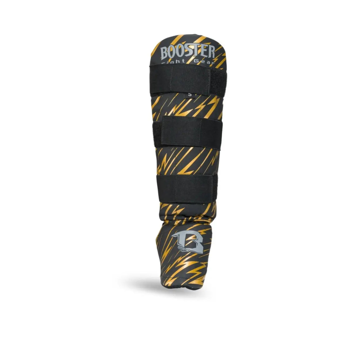 Kickboksscheenbeschermer van Booster in zwart met gouden bliksemprint, zilverkleurig logo op de voet en drie stevige klittenbandsluitingen.









