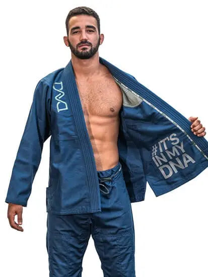 Man in blauwe BJJ-gi met open jas waarop de tekst ‘#IT'S IN MY DNA’ zichtbaar is, staand op een witte achtergrond.