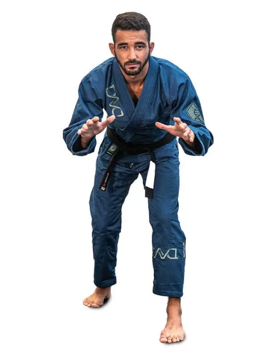 Man in blauwe BJJ-gi met zwarte band staat in lage vechthouding op een witte achtergrond.