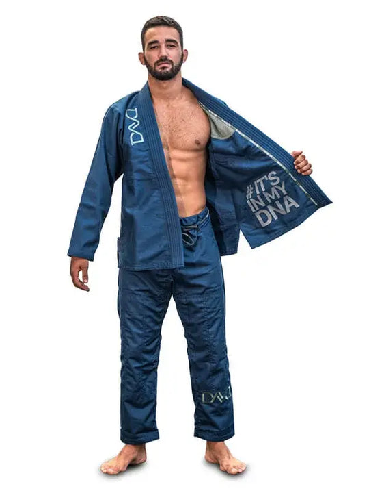 Man in blauwe BJJ-gi met open jas en zichtbare tekst ‘#IT'S IN MY DNA’, staand op een witte achtergrond.