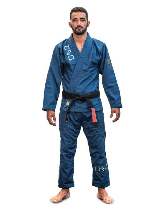 Man in blauwe BJJ-gi met zwarte band staat rechtop en kijkt recht vooruit op een witte achtergrond.