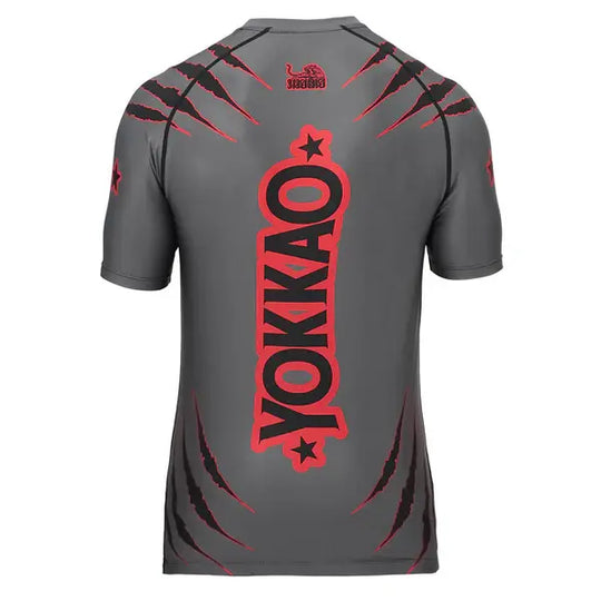 Achteraanzicht van het Yokkao compression shirt in grijs met rood en zwart design. Op de rug staat een groot verticaal Yokkao-logo in rood met zwarte omlijning, gecombineerd met rode krasdetails. De nauwsluitende pasvorm en korte mouwen bieden optimale ondersteuning en bewegingsvrijheid tijdens intensieve trainingen.