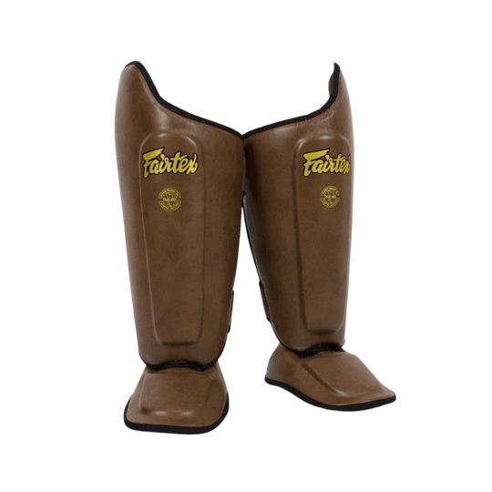 Een paar staande bruine scheenbeschermers met Fairtex-logo in goudkleurige letters.