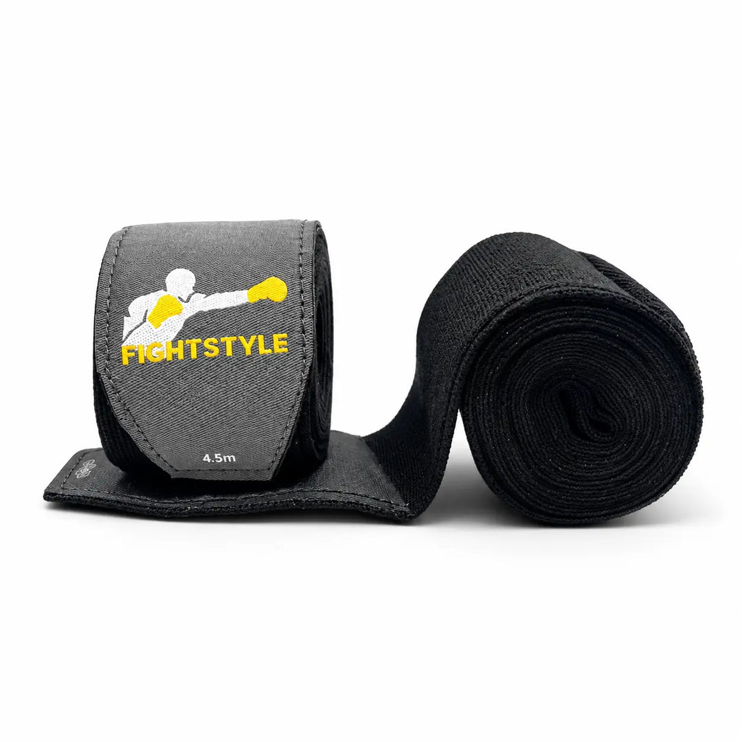 Zwarte Fightstyle bokswraps van 4,5 meter met grijs klittenbandlabel en geel-wit logo. De elastische stof ondersteunt de handen en polsen tijdens boksen, kickboksen en intensieve stoottrainingen.