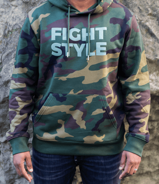 Close-up van de Fightstyle hoodie in camouflagegroen, gedragen door een model met jeans. De hoodie heeft een militair design, een zacht fleece-interieur en het herkenbare Fightstyle-logo op de borst — een statement piece voor fighters met stijl.