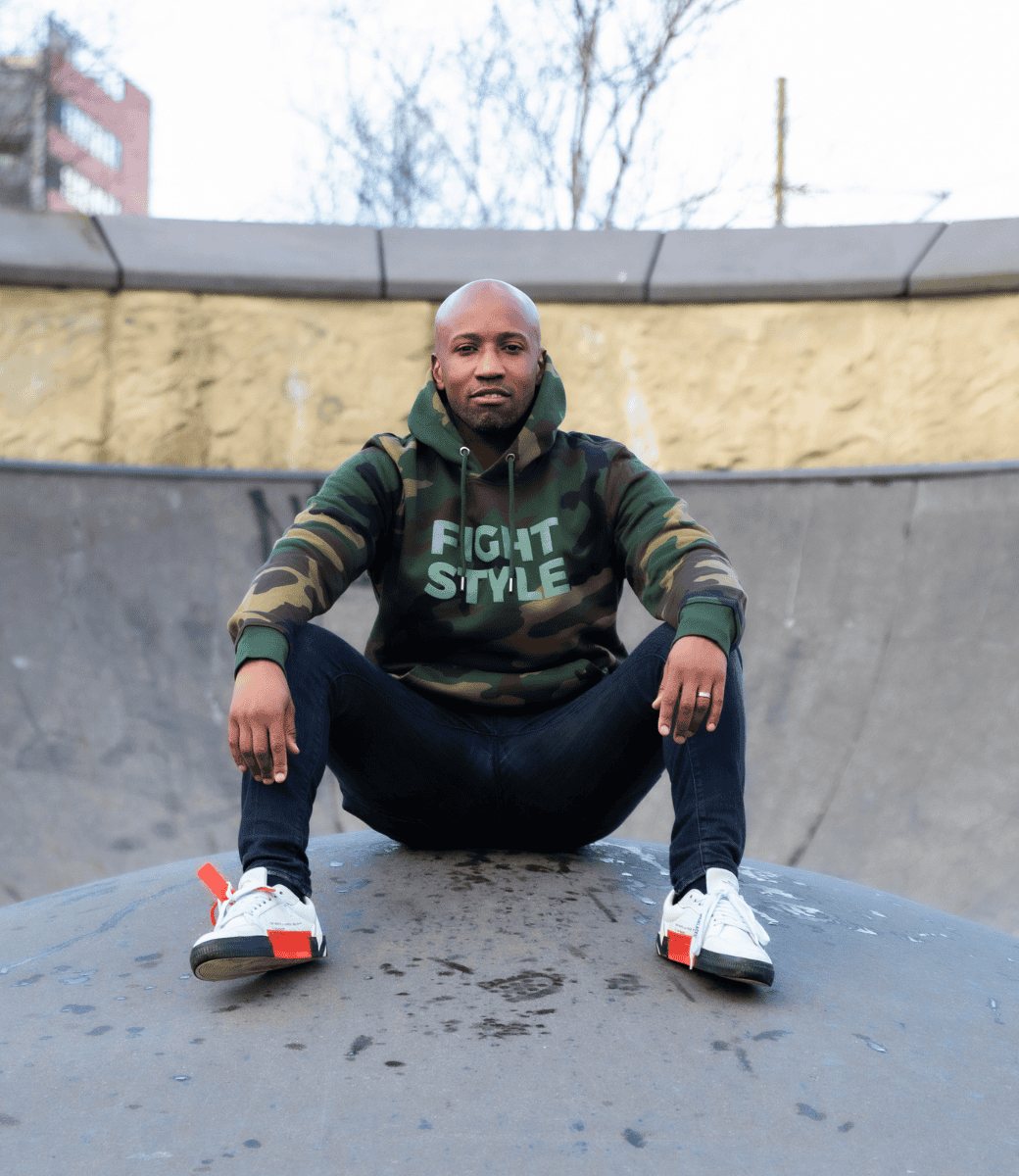 Mannelijk model draagt de Fightstyle hoodie Military Green met camouflagepatroon, zittend in een urban skatepark. De hoodie heeft een stoere streetwear-look met een zachte fleecevoering en een opvallend logo op de borst, ideaal voor sport of casual stijl.