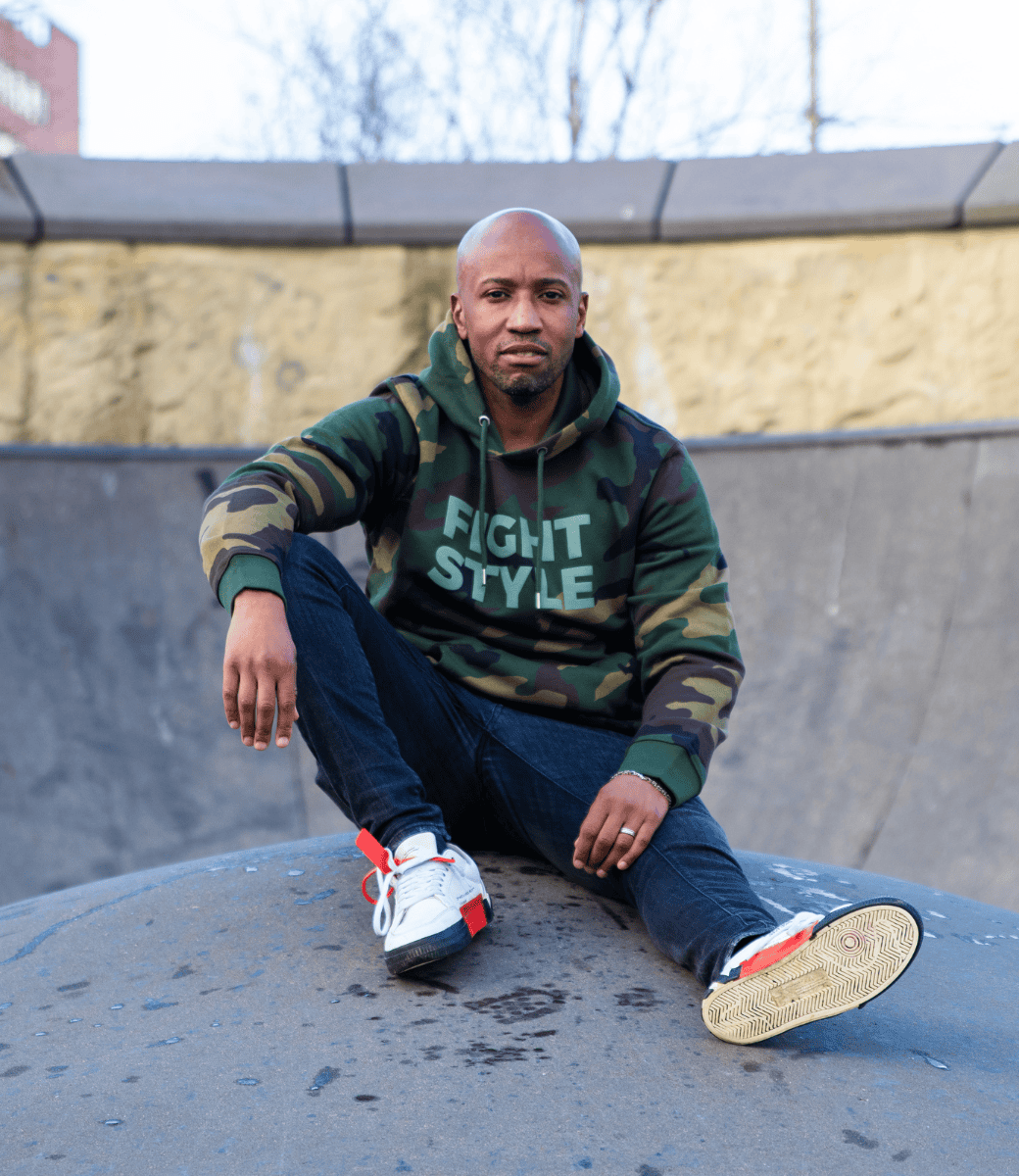 Man draagt de Fightstyle hoodie Military Green met camouflageprint, zittend in een skatepark. De hoodie heeft een stoere urban uitstraling met zachte voering, ideaal voor sportieve en casual outfits met karakter.