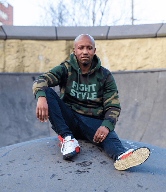 Man draagt de Fightstyle hoodie Military Green met camouflageprint, zittend in een skatepark. De hoodie heeft een stoere urban uitstraling met zachte voering, ideaal voor sportieve en casual outfits met karakter.