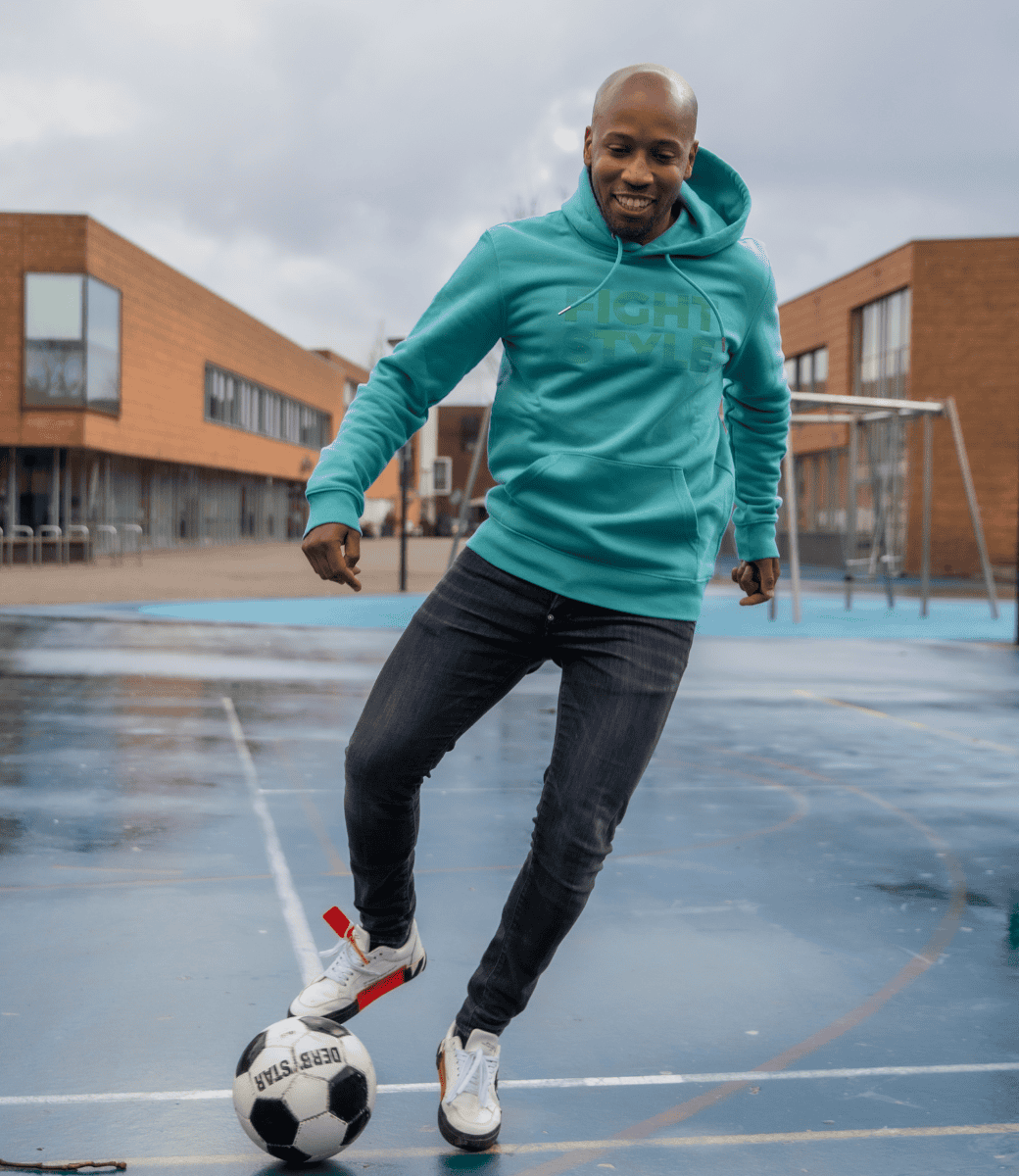 Man draagt de Fightstyle hoodie Teal Monstera terwijl hij voetbalt op een schoolplein. De hoodie combineert een sportieve look met comfort en stijl, perfect voor actieve dagen en casual momenten.