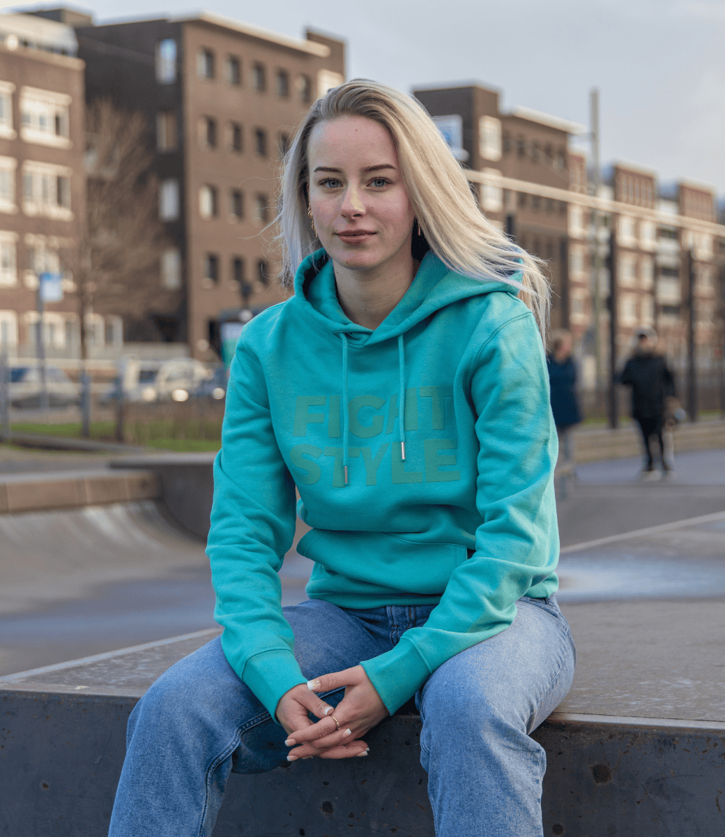 Vrouw draagt de Fightstyle hoodie Teal Monstera in een stadsomgeving. De hoodie heeft een frisse turquoise kleur, relaxed fit en subtiel logo, perfect voor zowel casual dagen als sportieve momenten.