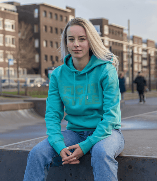 Vrouw draagt de Fightstyle hoodie Teal Monstera in een stadsomgeving. De hoodie heeft een frisse turquoise kleur, relaxed fit en subtiel logo, perfect voor zowel casual dagen als sportieve momenten.