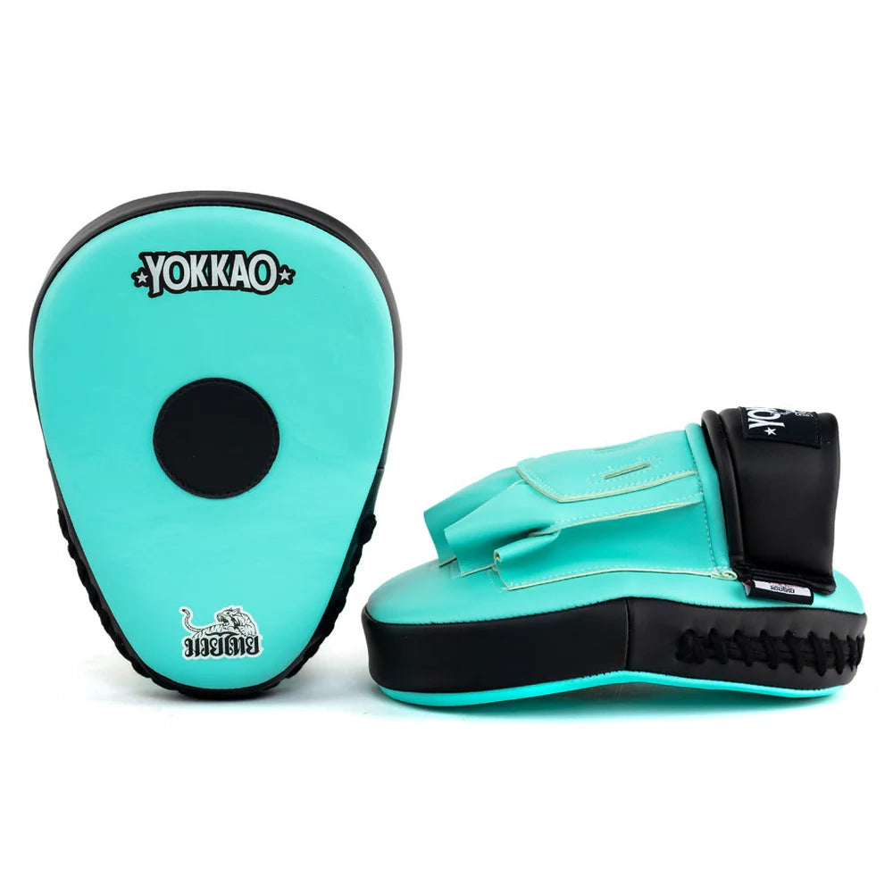 Yokkao focus mitts in turquoise en zwart met wit YOKKAO-logo, gefotografeerd tegen een lichte achtergrond. Links de voorzijde met zwart rond doelvlak en Muay Thai-embleem, rechts de zijkant met handinvoer, polsband en zwarte veters langs de onderrand. Geschikt voor nauwkeurige en krachtige padstraining tijdens boksen en Muay Thai in de gym.