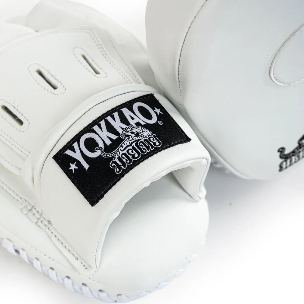 Close-up van Yokkao focus mitt in wit met zwarte logo patch op de polsband. De ventilatieopeningen en fijne stiksels zijn zichtbaar op de witte buitenzijde. Detailopname van de afwerking voor bokstraining en Muay Thai padsessies in de gym.